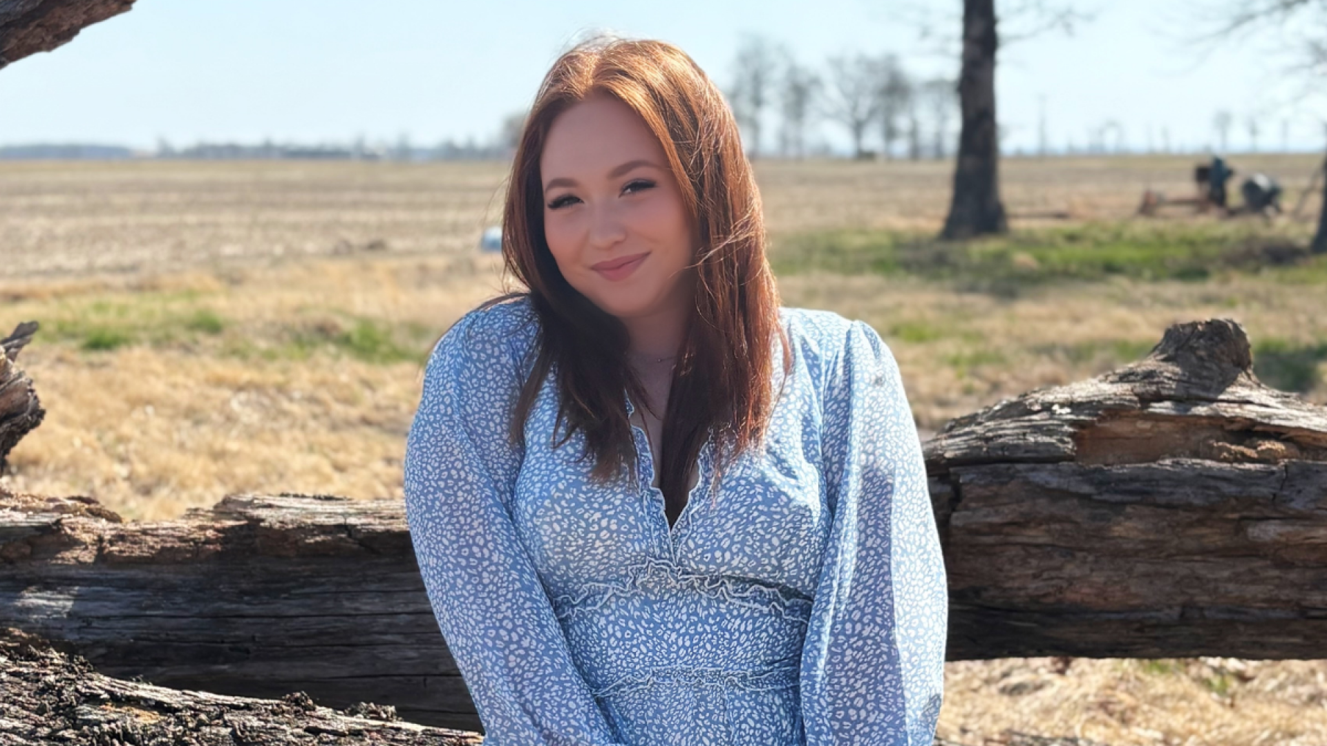 A young woman with long red hair, wearing a light blue patterned top, smiles while leaning against a fallen tree in a rural field.